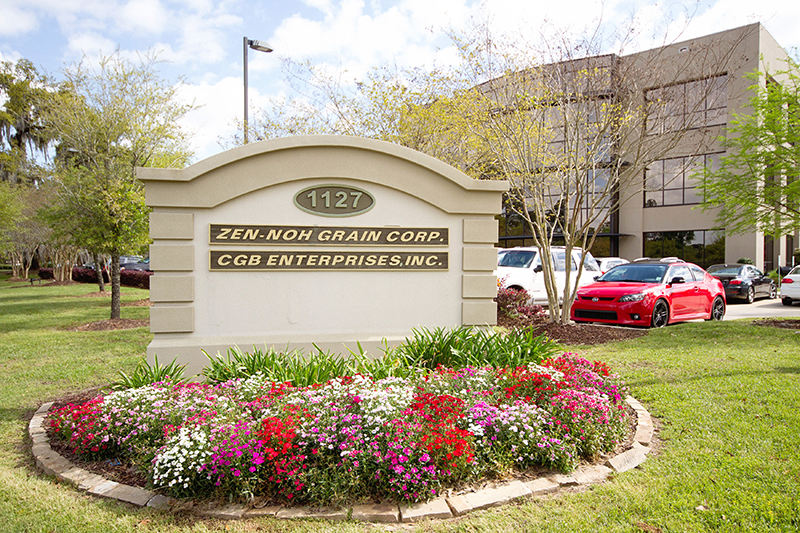 photo of a stucco sign in front of a large building that reads Zen-Noh Grain Corp and CGB Enterprises, Inc. The sign is surrounded by trees and flowers in differen colors such as white, red, and pink.