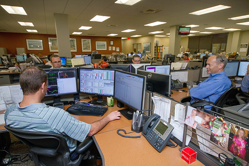 photo of inside the CGB office of employees working at their desks. One shown has 3 black computer monitors.